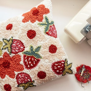 May include: A cream-colored pillow with a hand-stitched design featuring red strawberries, green leaves, and orange flowers. The pillow is made with a fluffy, textured fabric.