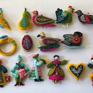 May include: A collection of thirteen handcrafted felt ornaments featuring various shapes including birds, a pear, a ring, an egg, a heart, and several figures in colourful clothing with intricate embroidery, decorative stitching, and small pins.