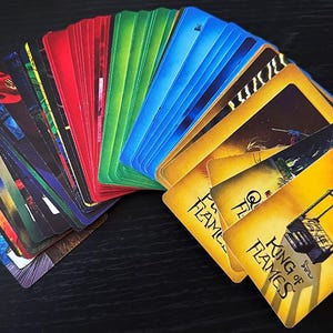 Op de afbeelding: Een stapel kleurrijke speelkaarten uitgespreid op een donkere ondergrond. De kaarten tonen verschillende afbeeldingen en tekst, met de nadruk op de kaart "King of Flames". De kaarten zijn gerangschikt in een regenboogspectrum.