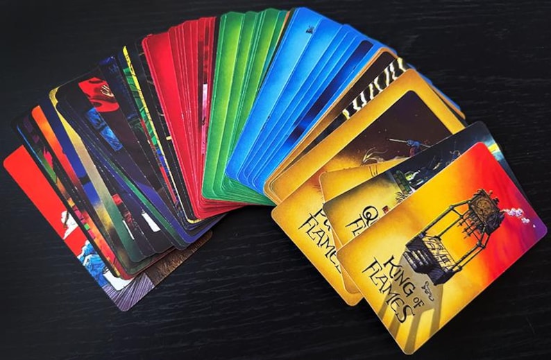 Op de afbeelding: Een stapel kleurrijke speelkaarten uitgespreid op een donkere ondergrond. De kaarten tonen verschillende afbeeldingen en tekst, met de nadruk op de kaart "King of Flames". De kaarten zijn gerangschikt in een regenboogspectrum.