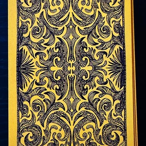 Op de afbeelding: Een tarotkaart met een gele achtergrond en een zwart, sierlijk ontwerp. Het ontwerp is symmetrisch en bevat wervelende patronen en bloemmotieven. De randen van de kaart zijn ook geel, wat een samenhangende uitstraling cre&euml;ert.