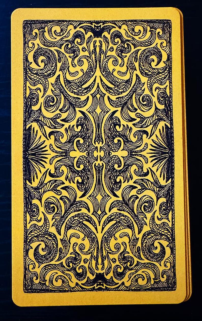 Op de afbeelding: Een tarotkaart met een gele achtergrond en een zwart, sierlijk ontwerp. Het ontwerp is symmetrisch en bevat wervelende patronen en bloemmotieven. De randen van de kaart zijn ook geel, wat een samenhangende uitstraling cre&euml;ert.