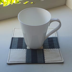 May include: A white ceramic mug with a curved handle rests on a square patchwork coaster. The coaster has a grid pattern of white, grey, and dark grey fabric squares, with visible stitching. The mug is empty.