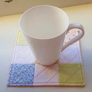 May include: A white ceramic mug sits on a handmade patchwork coaster. The coaster is made of pastel pink, green, and blue floral fabric squares. The mug has a curved handle and a simple design. The coaster is approximately 15 cm square.