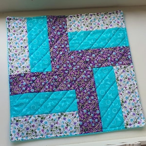May include: Square quilted fabric item with a patchwork design. The design features turquoise, purple floral, and white floral patterned fabric sections. The turquoise sections have a quilted diamond pattern. The item has a turquoise border.