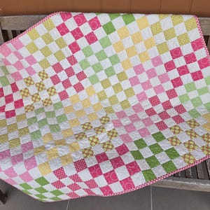 Peut inclure: Une courtepointe en patchwork composée d'une grille de carrés dans des tons de rose, jaune et vert lime. Les carrés de tissu présentent divers motifs, notamment de petits points et des fleurs. La bordure est à carreaux roses et blancs.
