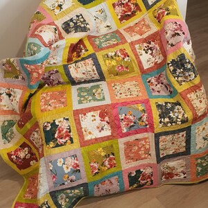 May include: A floral patchwork quilt draped over a chair. The quilt displays squares of various floral patterns in pink, yellow, green, and blue. The quilt is made of soft fabric. The quilt's measurements are in centimetres.