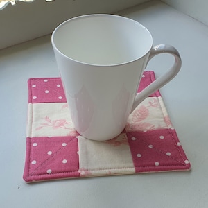 May include: A white ceramic mug sits on a handmade square coaster. The coaster is a patchwork design with pink polka dot and floral fabric squares. The mug has a curved handle and a simple, elegant design.