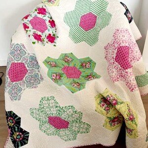 May include: A floral patterned quilt. The quilt features large, hexagon-shaped flowers in pink, green, and white. The flowers are set against a white background, with a pink border. The quilt is draped over a chair, showcasing the patchwork design.