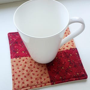 May include: A white ceramic mug rests on a quilted square coaster. The coaster is made of red and beige fabric squares with a floral design. The coaster is diamond-quilted and has a white border.