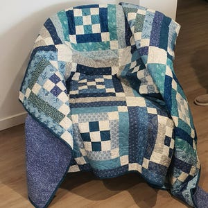 May include: A patchwork quilt in shades of blue, white, and teal, draped over a chair. The quilt features a variety of patterns, including squares and rectangles, with floral and geometric details. The quilt is made of soft fabric and is a handmade item.