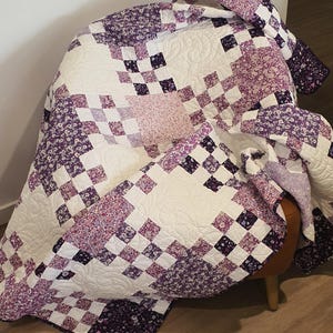 May include: A handmade quilt with a patchwork design in shades of purple, white, and floral patterns. The quilt is draped over a chair, showcasing its intricate stitching and textures. The quilt's dimensions are not visible.