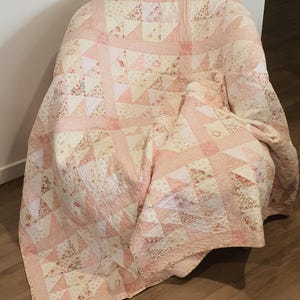 May include: A handmade quilt draped over a chair. The quilt features a patchwork design with pink and cream-coloured fabrics. The quilt has a floral pattern and a geometric design.
