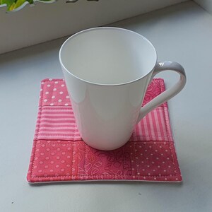 May include: A white ceramic mug with a grey handle sits on a square, quilted coaster. The coaster is made of pink fabric squares with patterns like polka dots and stripes. The mug is placed centrally on the coaster.