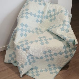 May include: A handmade quilt draped over a chair. The quilt features a repeating pattern of light blue and white squares. The quilt is made of soft fabric and has a quilted design with floral patterns.