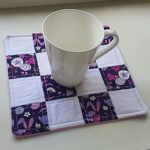 May include: A white ceramic mug sits on a quilted square coaster. The coaster has a patchwork design with alternating white squares and squares with a floral pattern in purple, pink, and white. The coaster has a pink border.