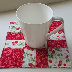May include: A white ceramic mug rests on a quilted square coaster. The coaster has a patchwork design with red floral and strawberry patterns. The mug has a curved handle and a smooth, glossy finish. The coaster is approximately 15 cm square.