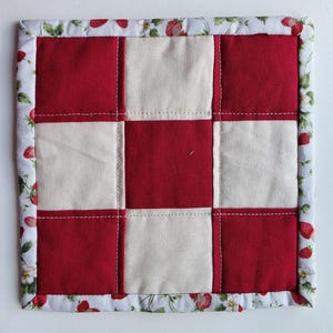 May include: A square quilted potholder featuring a checkerboard pattern of solid red and off-white fabric squares. The piece is finished with a white border fabric decorated with a repeating pattern of red strawberries and green leaves.