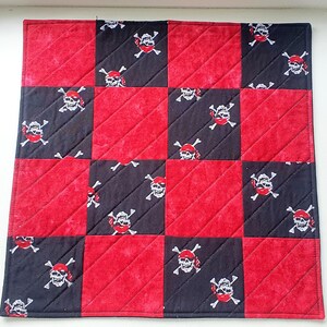 May include: A square, quilted fabric item with a checkerboard pattern of red and black squares. Each black square features a white skull and crossbones design with a red bandana. The item has a red border and diagonal stitching.