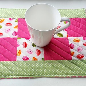 May include: A white mug sits on a handmade quilted placemat. The placemat has a patchwork design with pink, white floral, and green polka dot fabric. The mug has a grey handle.