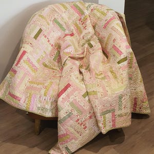 May include: A patchwork quilt in pink, green, and cream hues. The quilt's design is made up of rectangular fabric pieces. The quilt is draped over a wooden chair. The quilt is a handmade item.