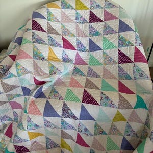 May include: A patchwork quilt featuring a repeating pattern of half-square triangles in various colours including yellow, pink, purple, teal, and white. The fabric pieces display diverse floral, polka dot, and geometric prints with visible quilting stitches.