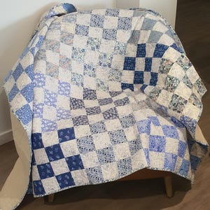May include: A blue and white patchwork quilt draped over a chair. The quilt has a checkerboard pattern with squares of varying blue floral and solid designs. The quilt is made of soft fabric and has a light beige backing.