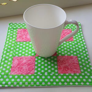 May include: A plain white ceramic mug sits on a square quilted fabric coaster. The coaster features a patchwork design alternating between bright green fabric with white polka dots and pink fabric with a subtle tonal swirling pattern.