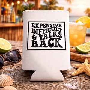 May include: A white can cooler with the text "EXPENSIVE DIFFICULT & TALKS BACK" in a black, bold font. The cooler sits on a wooden surface with a cocktail, lime, sunglasses, and seashells.