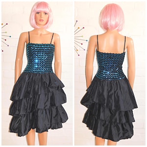 Könnte beinhalten: Ein ärmelloses Kleid mit einem eng anliegenden Oberteil, das mit schimmernden türkisfarbenen Pailletten und dünnen schwarzen Trägern versehen ist. Der Rock besteht aus mehreren Lagen schwarzem Stoff mit einem voluminösen Rüschendesign.