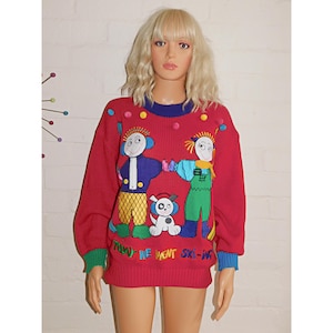 May include: A red long-sleeve knit jumper featuring a colourful graphic of two cartoon figures and a dog. The text "TODAY WE WENT SKI-ING" is embroidered at the bottom. The jumper has a blue crew neckline, green and blue cuffs, and small pom-poms.