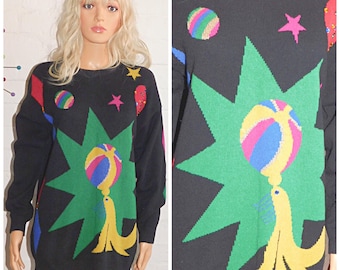 May include: Black sweater with a colourful circus-themed design. The sweater features a green starburst, a yellow seal balancing a striped ball, and various geometric shapes and stars in bright colours.