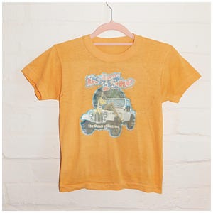 May include: A mustard yellow short-sleeve t-shirt featuring a graphic print of a white Jeep with a person sitting on the bonnet. The text Daisy Duke and The Dukes of Hazzard is printed in blue and red lettering above and below the vehicle graphic.