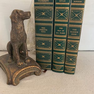 May include: A bronze dog bookend next to three vintage books with green covers and gold lettering. The books are titled "The Forsyte Saga", "The Way of All Flesh", and "The Razor's Edge". The bookend is on a decorative base.