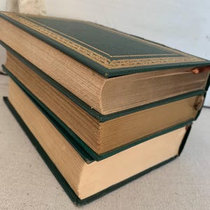 May include: Three antique books stacked. The books have dark green covers with gold trim and cream-coloured pages. The books are angled, showing their spines and edges. The books are likely vintage or second-hand.