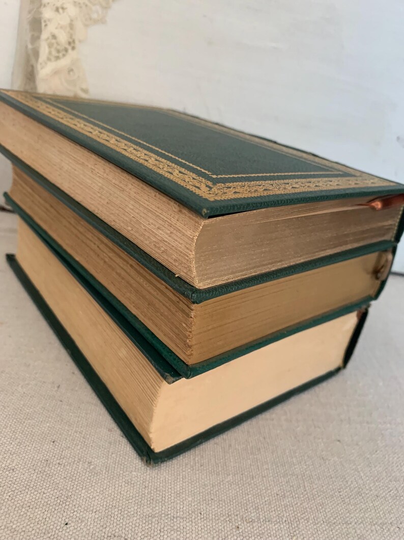 May include: Three antique books stacked. The books have dark green covers with gold trim and cream-coloured pages. The books are angled, showing their spines and edges. The books are likely vintage or second-hand.