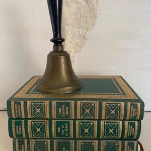 May include: A brass bell with a black handle sits on three green hardback books. The books have gold lettering and decorative details. Titles include "The Forsyte Saga", "The Way of All Flesh", and "The Razor's Edge".