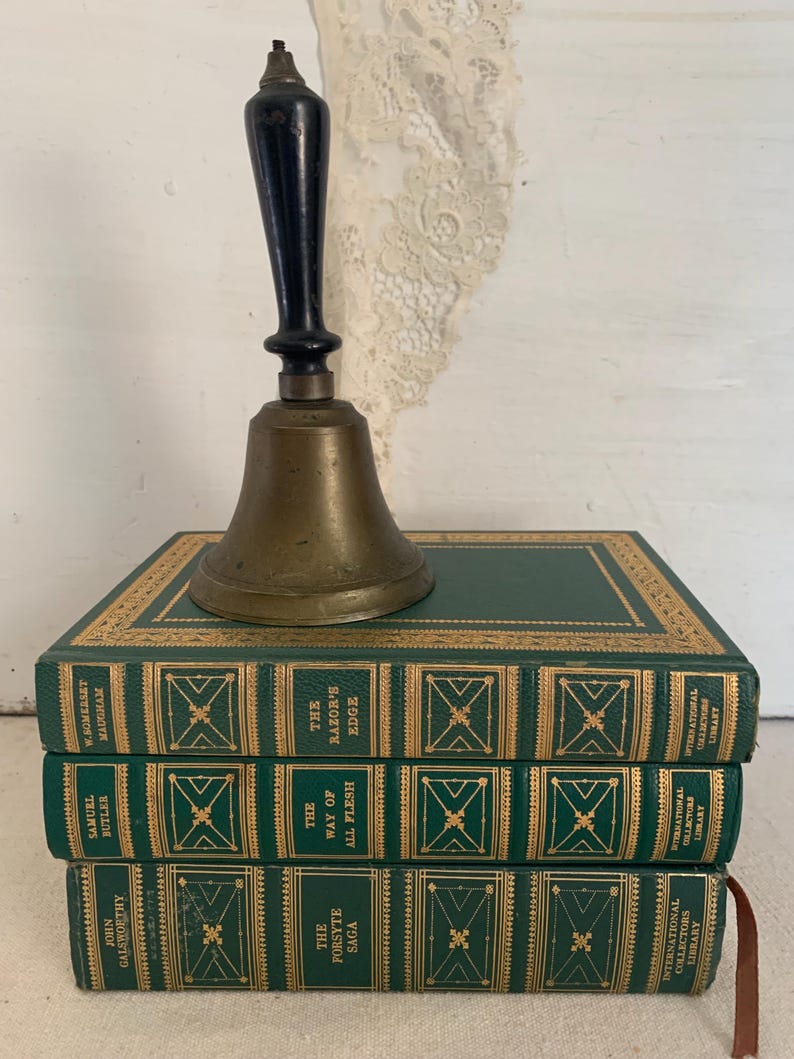 May include: A brass bell with a black handle sits on three green hardback books. The books have gold lettering and decorative details. Titles include "The Forsyte Saga", "The Way of All Flesh", and "The Razor's Edge".