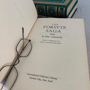May include: An open book titled "The Forsyte Saga" by John Galsworthy, with a preface by Ada Galsworthy. The book is from the International Collectors Library, Garden City, New York. A pair of spectacles rests on the page, with green books in the background.