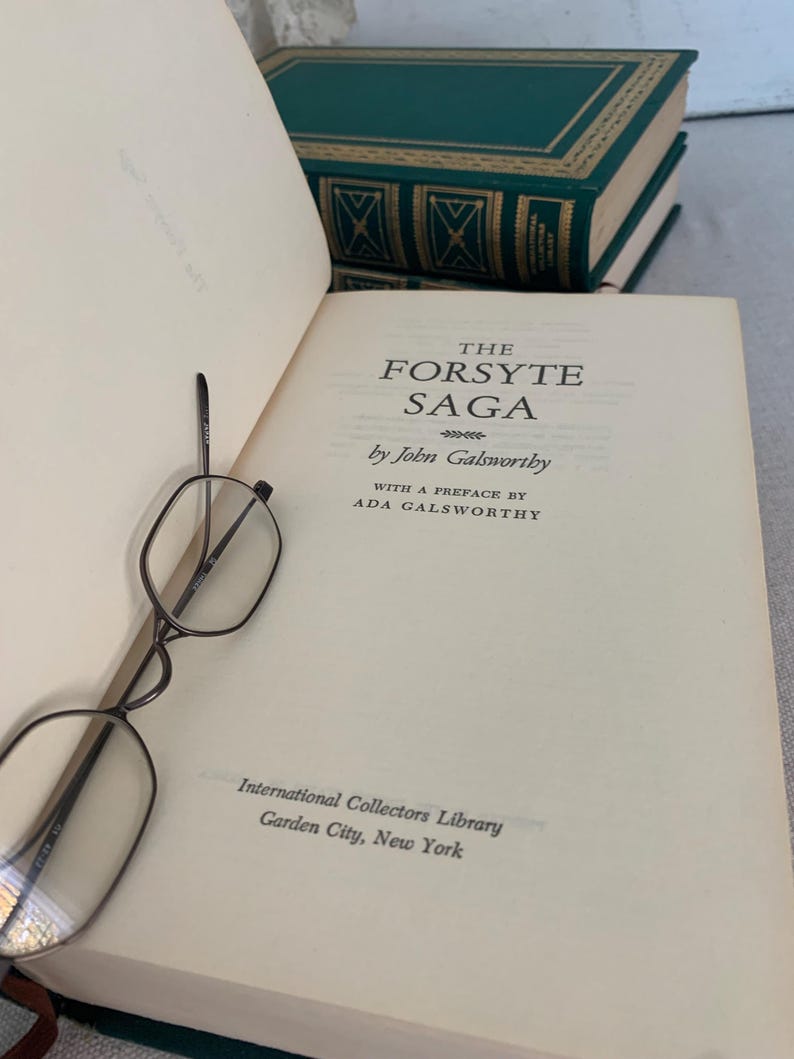 May include: An open book titled "The Forsyte Saga" by John Galsworthy, with a preface by Ada Galsworthy. The book is from the International Collectors Library, Garden City, New York. A pair of spectacles rests on the page, with green books in the background.