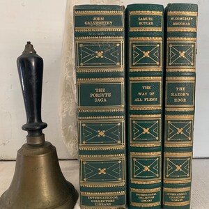 May include: A brass bell with a black handle is positioned next to three green hardcover books with gold detailing. The books feature titles such as "The Forsyte Saga", "The Way of All Flesh", and "The Razor's Edge".