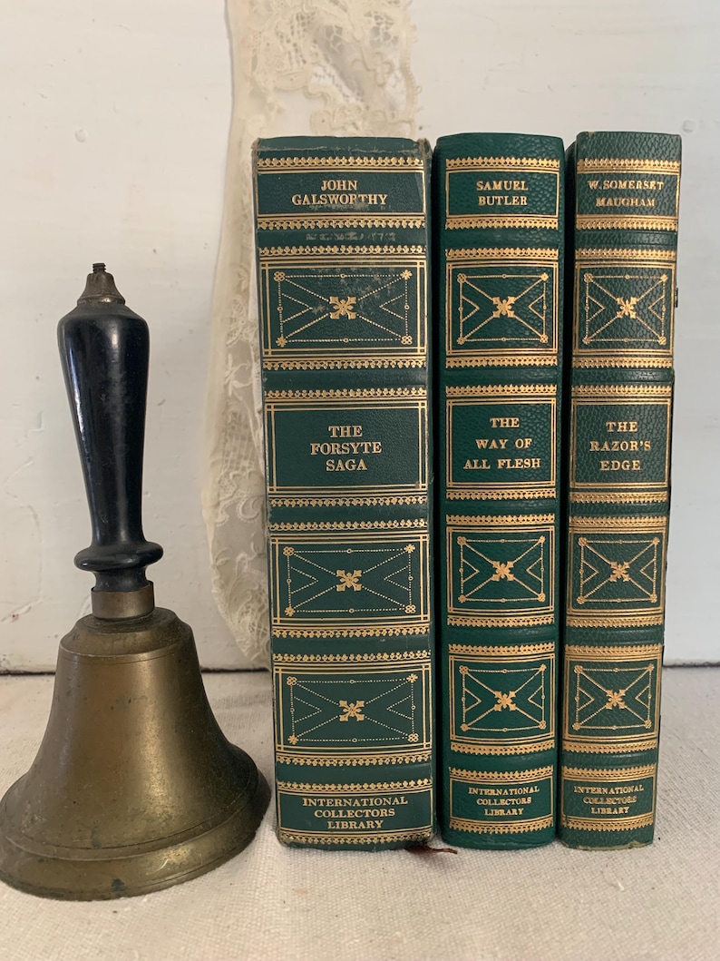 May include: A brass bell with a black handle is positioned next to three green hardcover books with gold detailing. The books feature titles such as "The Forsyte Saga", "The Way of All Flesh", and "The Razor's Edge".