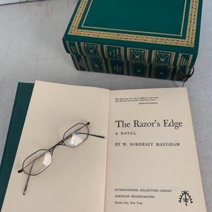 May include: An open book titled "The Razor's Edge" by W. Somerset Maugham, with eyeglasses resting on the page. A green book-shaped box with gold trim is in the background. The book is from the International Collectors Library.