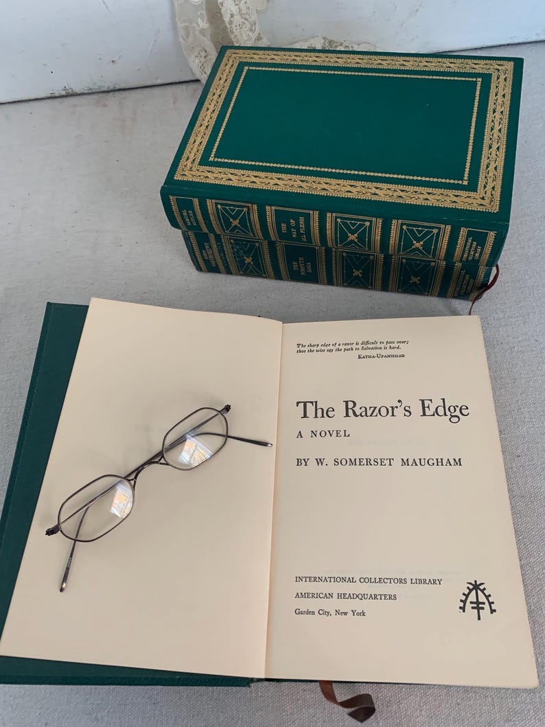 May include: An open book titled "The Razor's Edge" by W. Somerset Maugham, with eyeglasses resting on the page. A green book-shaped box with gold trim is in the background. The book is from the International Collectors Library.