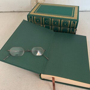May include: An open, green book with spectacles on the page. Two more green books with gold detailing are stacked behind it. A brown ribbon bookmark is visible. The books are likely hardback.