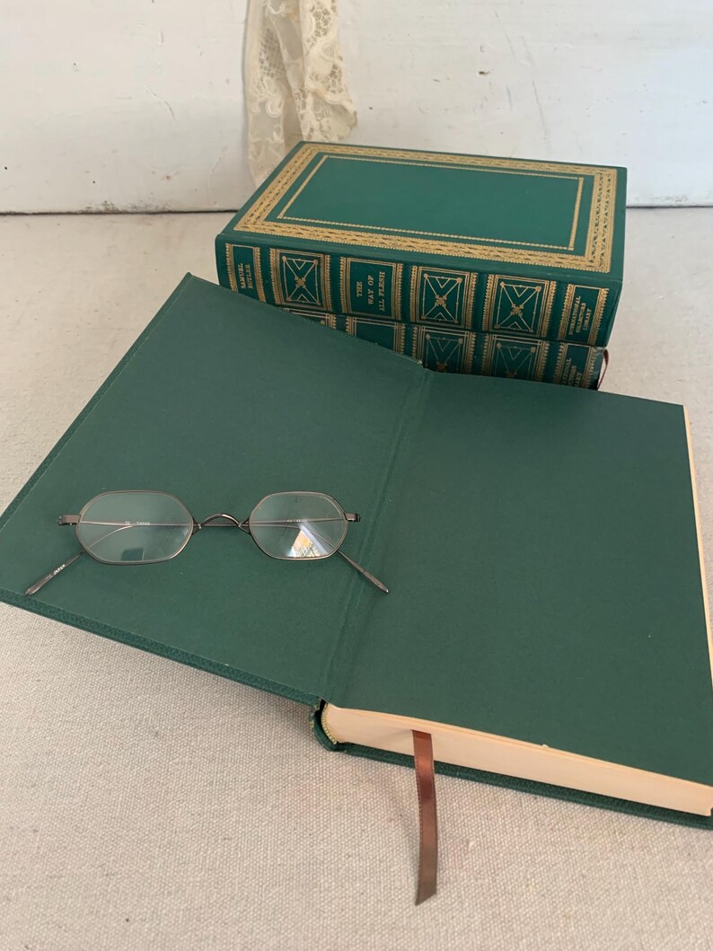 May include: An open, green book with spectacles on the page. Two more green books with gold detailing are stacked behind it. A brown ribbon bookmark is visible. The books are likely hardback.