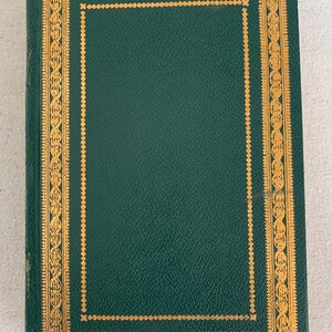 May include: A hardback book with a dark green cover and gold decorative border. The cover has a textured appearance. The gold border features an intricate design. A red ribbon bookmark is visible.