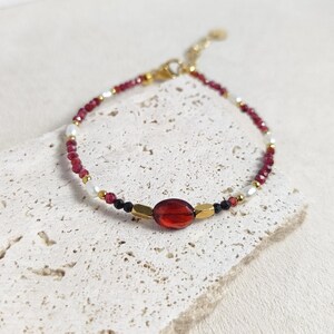 Könnte beinhalten: Ein zartes Armband mit roten, weißen und schwarzen Perlen, akzentuiert mit einem goldenen ovalen Edelstein. Das Armband liegt auf einer hellbeige texturierten Oberfläche.
