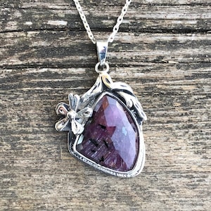 May include: A silver necklace with a purple gemstone pendant. The pendant is set in a silver frame with a floral design. The necklace is on a wooden background.
