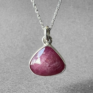 Peut inclure: Un collier en argent avec un pendentif en forme de triangle, orné d'une pierre précieuse rose foncé. Le pendentif est serti dans une lunette en argent et suspendu à une fine chaîne en argent. La pierre a une surface facettée.